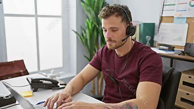 A young bearded man works attentively in an office, wearing a headset, in front of a laptop, exuding professionalism and focus