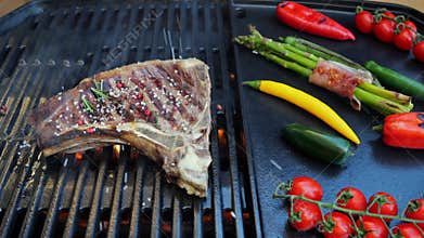 Beef T-Bone steak on grill sprinkle with spices, salt, peppercorns and rosemary, grilled vegetables. Slow motion. Close up
