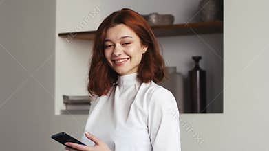 Portrait of beautiful caucasian woman tapping on smartphone, than smiling and looking to the camera while at home or