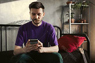 A man is using a smartphone while sitting at home with a guitar in the background