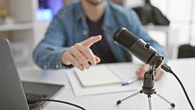 A focused man podcasting in a modern studio with a microphone, laptop, and pointing gesture, indicating interactive communication