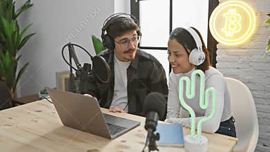 A man and woman with headphones podcasting in a modern studio with cryptocurrency neon signs