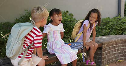 Diverse children sitting on bench with backpacks, boy coming and leaning, middle girl sharing story
