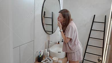 Woman examines skin on face while looking in bathroom mirror near sink