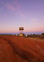 A water tower highlighted against a colorful sky and a red dirt foreground.