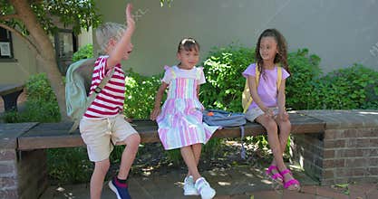Diverse children playing, watching boy fidgeting and giving high-fives on bench with backpacks