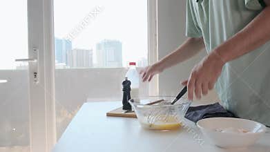 Man pouring milk and whisking beaten eggs in glass bowl in light kitchen during breakfast preparation. Cooking omelette