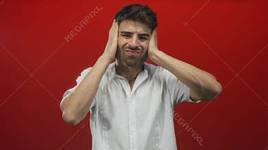 Hispanic man in a white shirt covering ears with hands in a red studio while grimacing in discomfort; noise pollution stress