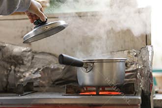 A woman's hand opens the lid of a pot of boiling water. Steam generated from boiling water shoots up to the top.