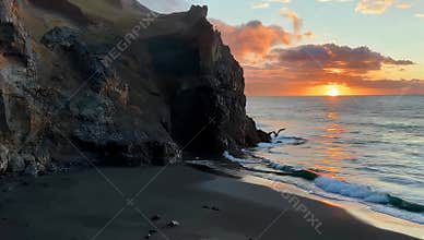 Dramatic sunset over a black sand beach with cliffs and ocean waves creating a scenic landscape