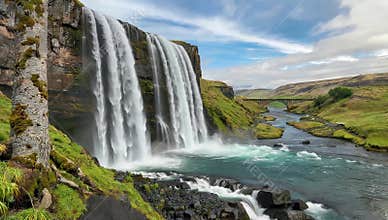 Majestic waterfall cascades down rocky cliffs into a vibrant river under a cloudy sky view