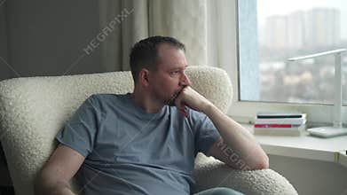 Man reflecting on his thoughts while sitting by a window in a cozy room during a cloudy afternoon