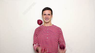 Young handsome guy in a striped sweater smiles broadly and juggles with three red apples.
