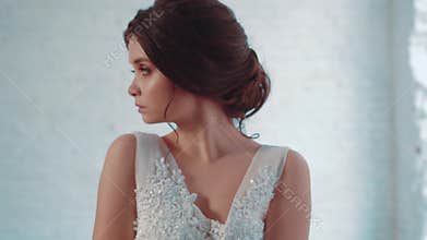 Mysterious young attractive brunette girl in a long, luxurious, white-faced dress stands in the studio, looks away