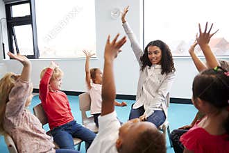 A class of infant school children sitting on chairs in a circle in the classroom, raising hands with their female teacher, close u