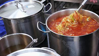 A cook in a restaurant mixes borsch in a large pan