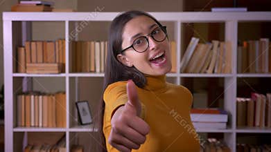 Happy and delighted brunette female teacher gestures thumb-up sign to show like and respect at the library.