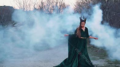 Wild witch in long free flying green emerald dress stands in thick white fog and raises her hands, a lady calls a
