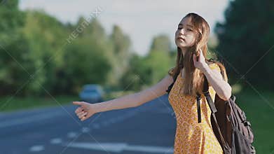 Young beautiful girl with long hair in a dress and a backpack on her back is catching a car on the freeway. She shows a