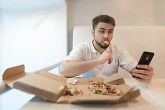 A beautiful and funny man eats a pizza out of the box and picks up his phone. Selfies with pizza