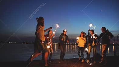 Group of young friends having a beach party. Friends dancing and celebrating with sparklers in twilight sunset