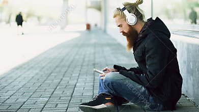 Young bearded hipster man with headphones sitting on road and using smartphone for listen to music and internet surfing