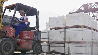 Forklift truck driver in a factory or warehouse driving between rows of shelving with stacks of boxes and packaging