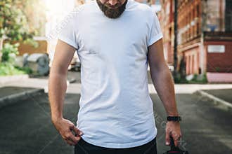 Front view. Young bearded millennial man dressed in white t-shirt is stands on city street. Mock up.
