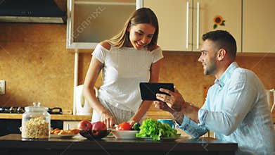 Attractive couple chatting in the kitchen early morning. Handsome man using tablet while his girlfriend cooking