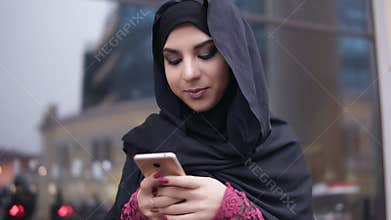 Closeup view of young attractive woman wearing hijab standing in the street, typing a message on her mobile phone