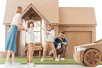 young family playing baseball together on yard of cardboard house