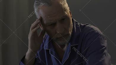 Elderly male sitting on bed in dark room, rubbing his temples, strong headache