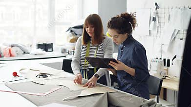 Attractive clothes designer entrepreneurs are outlining cutout with chalk, working with tablet and talking. Busy day at