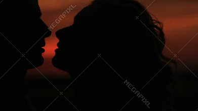 Close-up portrait of the charming silhouette couple gently kissing at the background of the red sky during the sunset.