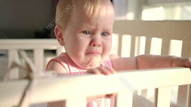 Sad baby crying in cot at home. Unhappy toddler standing in crib