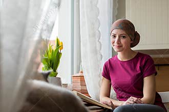 Young positive adult female cancer patient sitting in the kitchen by a window reading a book, smiling.