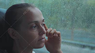 Portrait of young sad woman looking out the wet window, while travelling by bus.