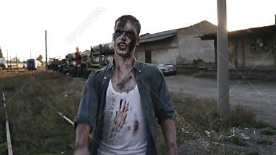 Creepy zombie man in bloody clothes walking by railway lines outdoors with an industrial abandoned place on the