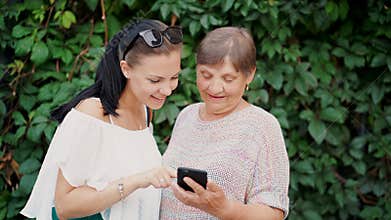 Granddaughter shows old grandmother something in smartphone, teaches her to handle with modern gadget and technology