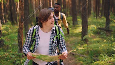 Smiling young man is looking at map and searching for right way in forest while multiethnic group of friends is
