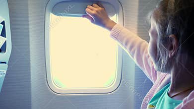 Close-up. Kid girl raises the porthole curtain in the airplane`s cabin, from there shines a bright light. girl looking