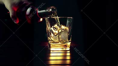 Barman pouring whiskey in the glass with ice cubes on wood table and black dark background, focus on ice cubes, whisky relax time