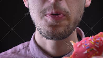 Close-up portrait of man biting and chewing delicious glazed pink donut with colorful chips and eyes on black background