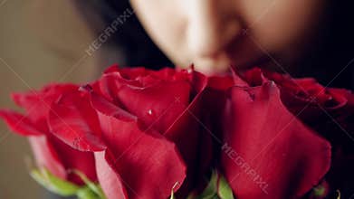 Beautiful girl sniffs a bouquet of red roses and then looks out the window. Closeup