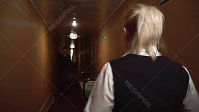 A maid rolls a trolley down the hall in the hotel and greets a man walking towards and talking on the cell phone.