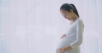 Beautiful asian pregnant woman looking through window and rubbing stomach at living room
