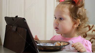 Child girl eating breakfast muesli with yogurt and loking a video on the tablet screen.