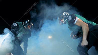Two American football players compete on a filed, side view.