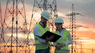 A man and a woman with a laptop are working beside power line towers. Engineers working on renewable energy development.