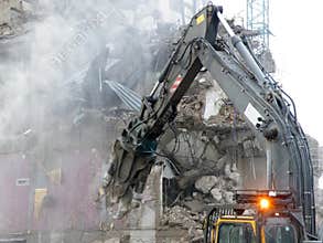 A mechanical digger with a pneumatic hammer tearing down and old building on a an urban development site with dust debris and